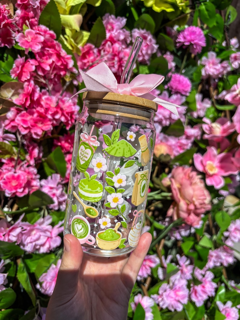 Puede incluir: Un vaso de vidrio transparente con tapa de bamb&uacute; y un lazo rosa. El vaso est&aacute; decorado con ilustraciones tem&aacute;ticas de matcha, que incluyen una taza de matcha, un batidor y un postre de matcha. Una pajita de vidrio transparente se inserta en el vaso.