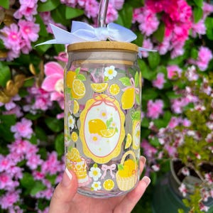 May include: A clear glass tumbler with a bamboo lid, a glass straw, and a white bow. The tumbler is decorated with yellow lemons, white flowers, and a yellow oval design. The tumbler is held in front of a background of pink flowers.