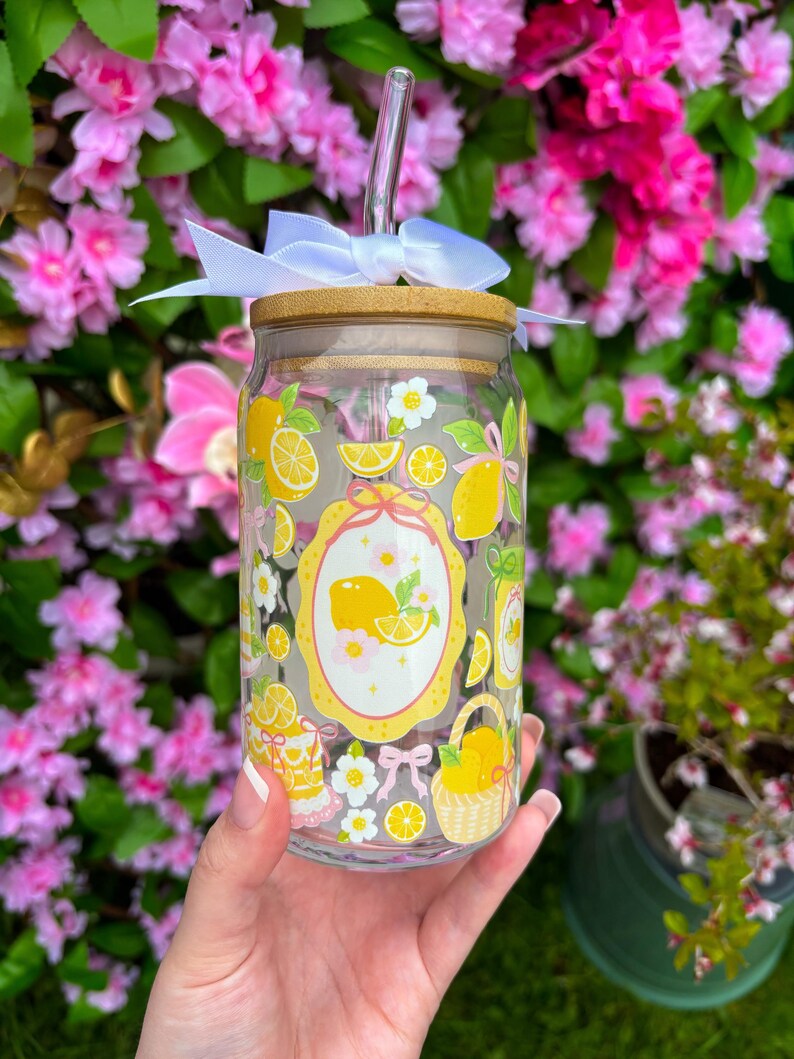 May include: A clear glass tumbler with a bamboo lid, a glass straw, and a white bow. The tumbler is decorated with yellow lemons, white flowers, and a yellow oval design. The tumbler is held in front of a background of pink flowers.