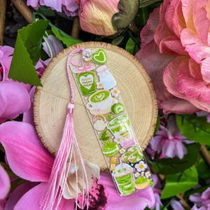 May include: A clear bookmark featuring matcha-themed illustrations, including matcha lattes, milk, and desserts. The bookmark has a pink tassel and is set against a backdrop of pink flowers and a wooden slice.