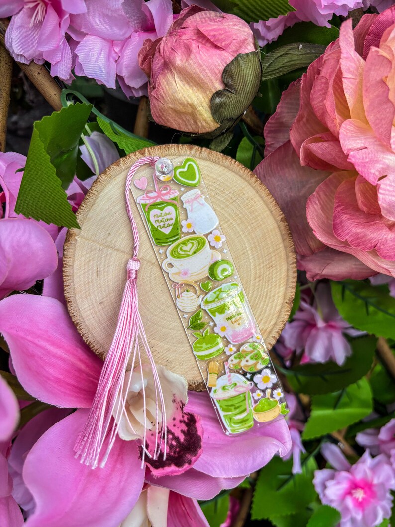 May include: A clear bookmark featuring matcha-themed illustrations, including matcha lattes, milk, and desserts. The bookmark has a pink tassel and is set against a backdrop of pink flowers and a wooden slice.