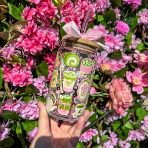 Puede incluir: Un vaso de vidrio transparente con tapa de bamb&uacute; y lazo rosa, con ilustraciones tem&aacute;ticas de matcha. Incluye una pajita transparente. El fondo es una vibrante exhibici&oacute;n de flores rosas y follaje verde.