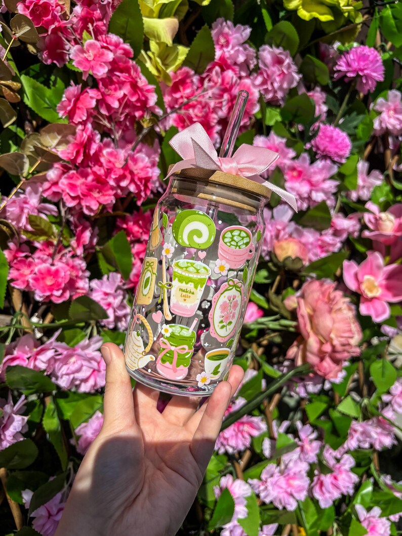 Puede incluir: Un vaso de vidrio transparente con tapa de bamb&uacute; y lazo rosa, con ilustraciones tem&aacute;ticas de matcha. Incluye una pajita transparente. El fondo es una vibrante exhibici&oacute;n de flores rosas y follaje verde.