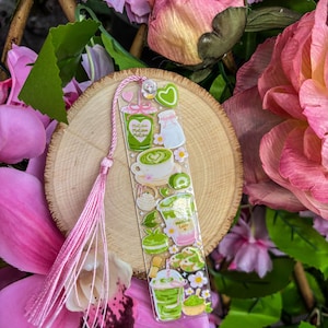 May include: A clear bookmark featuring matcha-themed illustrations in green, white, and pink. The bookmark includes images of matcha lattes, desserts, and a pink tassel. The background includes pink flowers and a wooden slice.