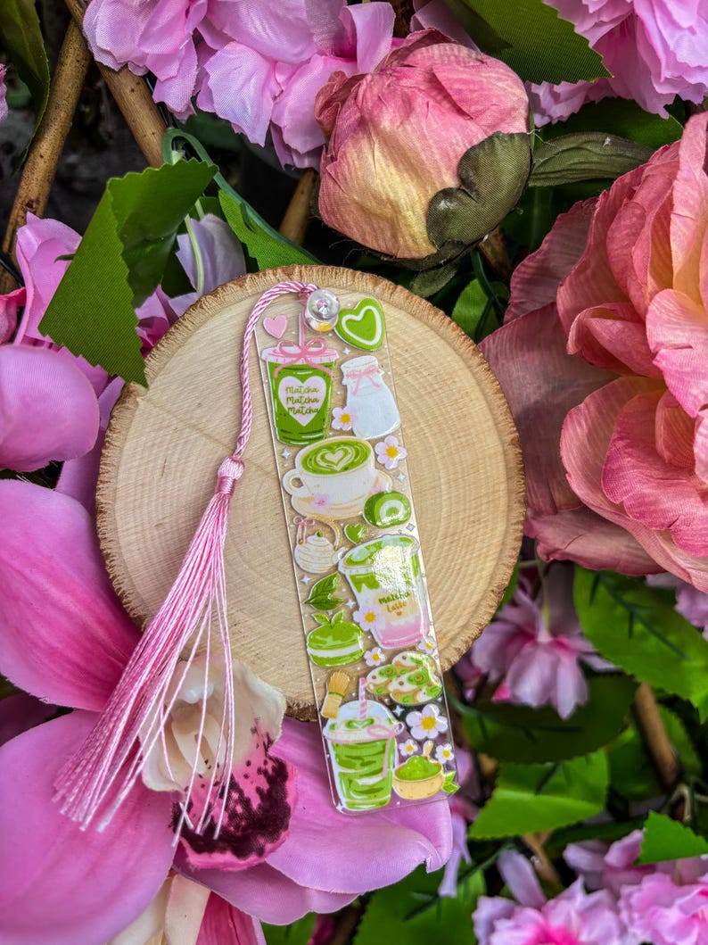 May include: A clear bookmark featuring matcha-themed illustrations in green, white, and pink. The bookmark includes images of matcha lattes, desserts, and a pink tassel. The background includes pink flowers and a wooden slice.