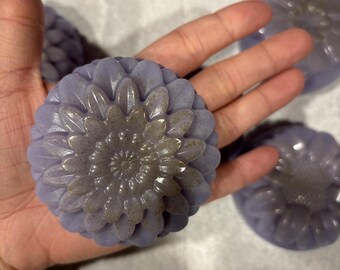 May include: Several lavender-coloured, flower-shaped soaps are shown in close-up. The soaps have a detailed floral design with a lighter centre. The soaps are on a white surface.