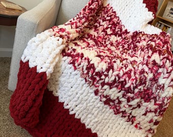 May include: A chunky knit blanket in a color block design. The blanket features alternating stripes of white, deep red, and a mix of red and white. The blanket is draped over a gray armchair.