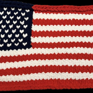 May include: A crocheted American flag in red, white, and blue. The flag features a blue square with white heart-shaped stars in the upper left corner and red and white stripes. The item is handmade.