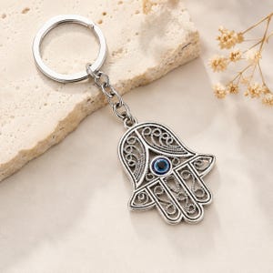May include: A silver-toned metal keychain featuring a Hamsa hand charm with intricate filigree cutouts and a central blue and black evil eye bead. The charm is attached to a standard circular split ring by a short silver-toned metal chain link.