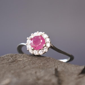 Può includere: Un anello d'argento con un grande rubino rosso rotondo e vibrante, circondato da un alone di pietre più piccole e chiare. L'anello è appoggiato su una superficie scura e strutturata, con uno sfondo grigio sfocato.