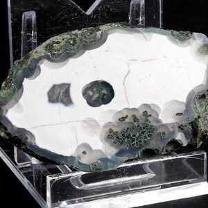 Puede incluir: Una rebanada de piedra de ágata pulida, que muestra un interior blanco y gris con un borde exterior verde oscuro y gris. La piedra tiene un agujero central y se exhibe sobre un soporte de acrílico transparente, resaltando sus patrones y colores naturales.