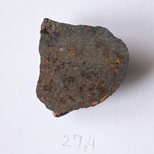 Pode incluir: Um fragmento de rocha cinza escuro, de forma irregular, com uma superfície áspera e texturizada. A rocha apresenta manchas de oxidação castanho-avermelhada e pequenas inclusões de cor clara. O número "27,4" está escrito a lápis no fundo branco.