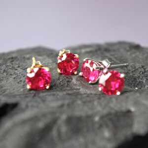 May include: Four stud earrings with vibrant red gemstones. Two pairs of earrings are visible, one with gold settings and the other with silver settings. The gemstones are round and faceted, reflecting light. The earrings are displayed on a dark, textured surface.