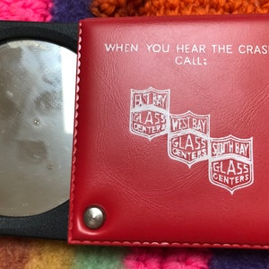 May include: A red leather compact mirror with a white inscription that reads "WHEN YOU HEAR THE CRASH! CALL: EAST BAY GLASS CENTERS, WEST BAY GLASS CENTERS, SOUTH BAY GLASS CENTERS".