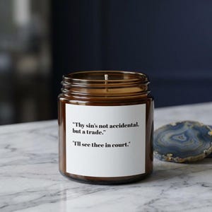 以下が含まれることがあります： 白いラベルが付いた茶色のガラス瓶のキャンドル。ラベルには「Thy sin's not accidental, but a trade.」と「I'll see thee in court.」と書かれています。キャンドルは大理石の表面に置かれています。