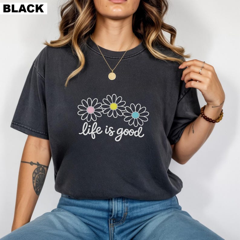 May include: Black t-shirt featuring a daisy graphic and the words "life is good" in white script. The daisies have pink, yellow, and blue centres. The shirt has a relaxed fit.