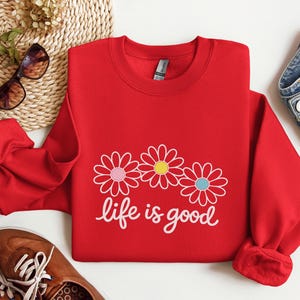 May include: A red sweatshirt featuring the text "life is good" in white script, with three daisy flower graphics. The flowers have pink, yellow, and blue centres. The sweatshirt is shown with blue jeans and brown shoes.