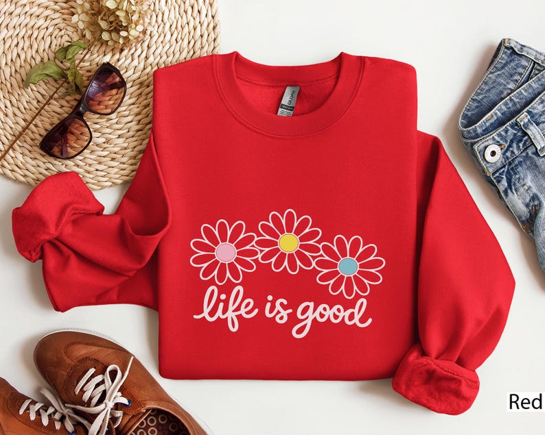 May include: A red sweatshirt featuring the text "life is good" in white script, with three daisy flower graphics. The flowers have pink, yellow, and blue centres. The sweatshirt is shown with blue jeans and brown shoes.
