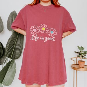 May include: A brick-red t-shirt featuring the text "life is good" in white script, with three daisy designs in pink, yellow, and blue. The top has short sleeves and a loose fit.