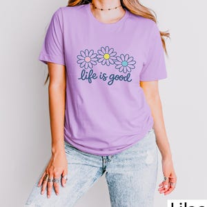 May include: A lilac t-shirt with a floral design. Three daisies with pink, yellow, and blue centres are above the cursive text "life is good". The shirt is a solid colour and has short sleeves.