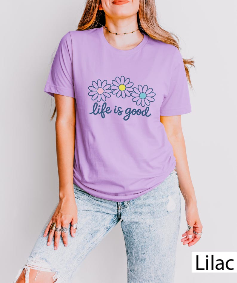 May include: A lilac t-shirt with a floral design. Three daisies with pink, yellow, and blue centres are above the cursive text "life is good". The shirt is a solid colour and has short sleeves.