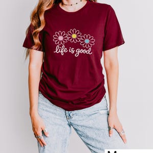 May include: A maroon t-shirt featuring the phrase "life is good" and three daisy illustrations with pink, yellow, and blue centres. The shirt is worn with light wash denim jeans.