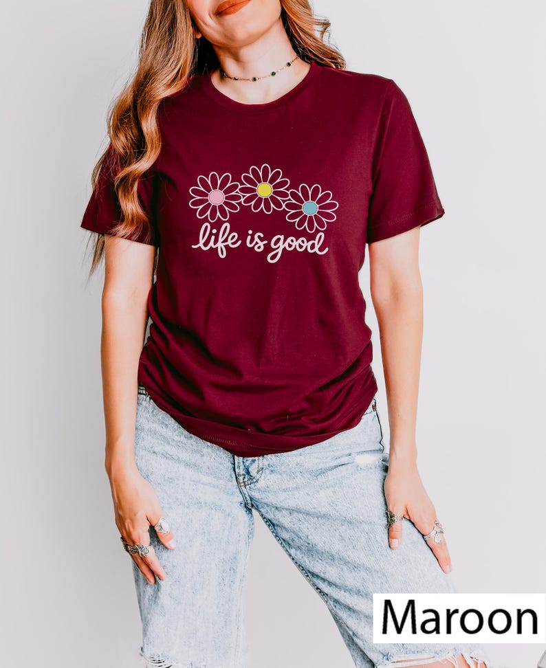 May include: A maroon t-shirt featuring the phrase "life is good" and three daisy illustrations with pink, yellow, and blue centres. The shirt is worn with light wash denim jeans.