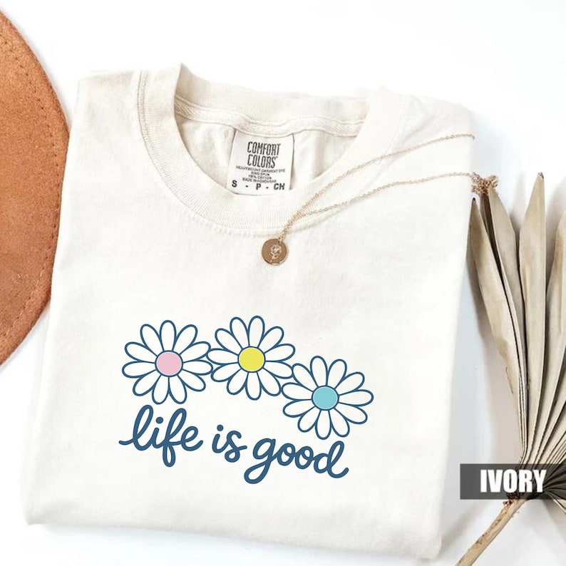 May include: Cream-coloured t-shirt featuring a daisy graphic and the navy blue phrase "life is good". The daisy flowers have pink, yellow, and blue centres. A gold necklace with a pendant is draped across the shirt.