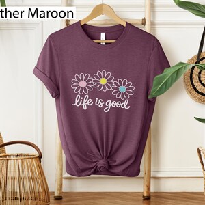 May include: Heather maroon t-shirt featuring three daisy flowers and the text "life is good" in white script. The daisies have pink, yellow, and blue centres. The shirt is displayed on a wooden hanger.