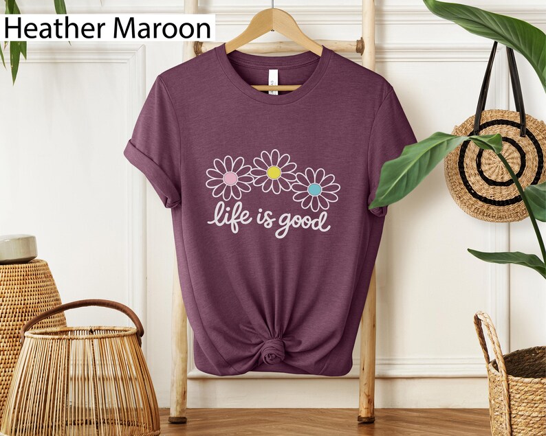 May include: Heather maroon t-shirt featuring three daisy flowers and the text "life is good" in white script. The daisies have pink, yellow, and blue centres. The shirt is displayed on a wooden hanger.