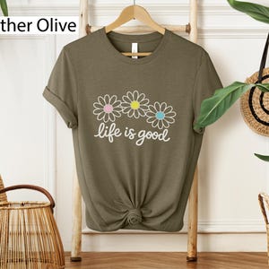 May include: Heather olive green t-shirt featuring a daisy graphic and the words "life is good". The daisy graphic has pink, yellow, and blue centres. The t-shirt is knotted at the bottom.