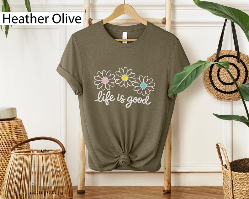 May include: Heather olive green t-shirt featuring a daisy graphic and the words "life is good". The daisy graphic has pink, yellow, and blue centres. The t-shirt is knotted at the bottom.