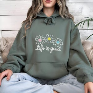 May include: Sage green hoodie with a front pocket and drawstring hood. The hoodie features a daisy graphic with pink, yellow, and blue centres, and the words "life is good" in white script. The hoodie is made of a soft material.