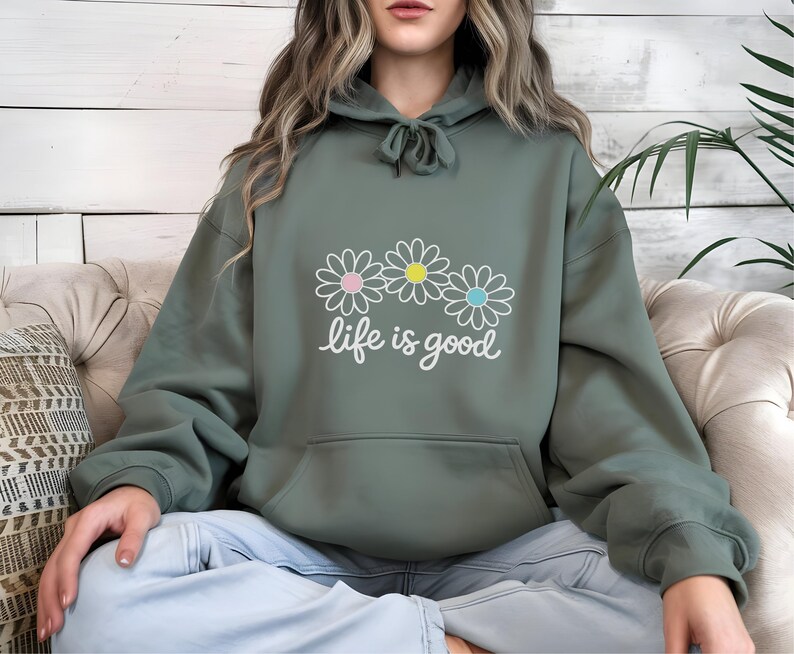 May include: Sage green hoodie with a front pocket and drawstring hood. The hoodie features a daisy graphic with pink, yellow, and blue centres, and the words "life is good" in white script. The hoodie is made of a soft material.