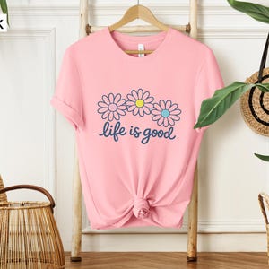 May include: A pink t-shirt featuring a daisy graphic and the words "life is good" in navy blue script. The daisy design has three flowers with blue, yellow, and pink accents. The shirt is displayed on a wooden rack.