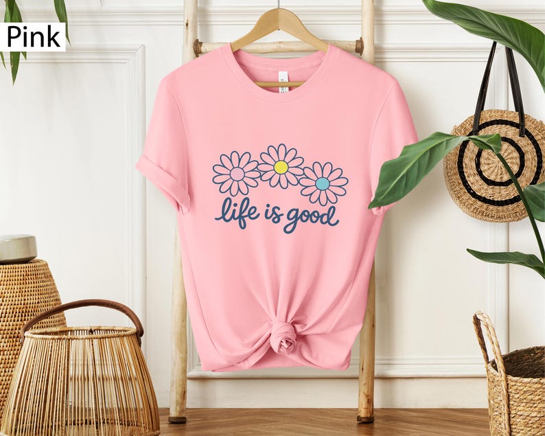 May include: A pink t-shirt featuring a daisy graphic and the words "life is good" in navy blue script. The daisy design has three flowers with blue, yellow, and pink accents. The shirt is displayed on a wooden rack.