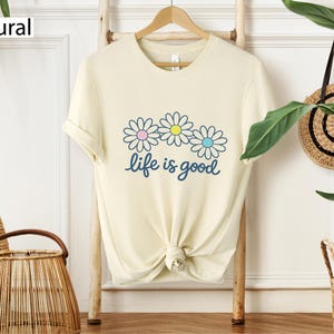 May include: Cream-coloured t-shirt featuring a daisy graphic and the phrase "life is good" in blue script. The daisy flowers have pink, yellow, and blue centres. The shirt is hanging on a wooden rack.