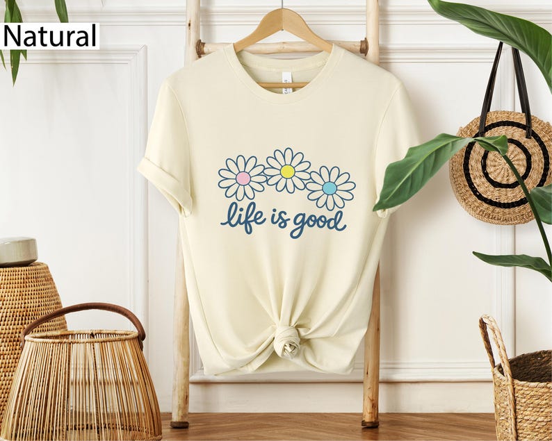 May include: Cream-coloured t-shirt featuring a daisy graphic and the phrase "life is good" in blue script. The daisy flowers have pink, yellow, and blue centres. The shirt is hanging on a wooden rack.