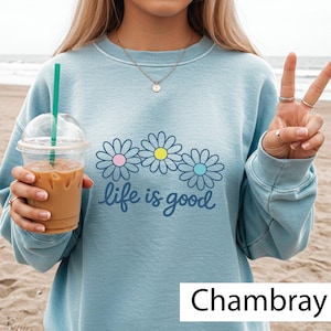 May include: Pale blue sweatshirt featuring a daisy design with pink, yellow, and blue centres, and the words "life is good." The sweatshirt is a chambray colour.
