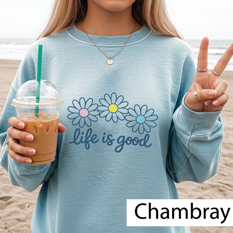 May include: Pale blue sweatshirt featuring a daisy design with pink, yellow, and blue centres, and the words "life is good." The sweatshirt is a chambray colour.