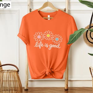 May include: Bright orange t-shirt featuring a floral design and the words "life is good" in white script. The t-shirt is knotted at the waist and hangs on a wooden hanger.
