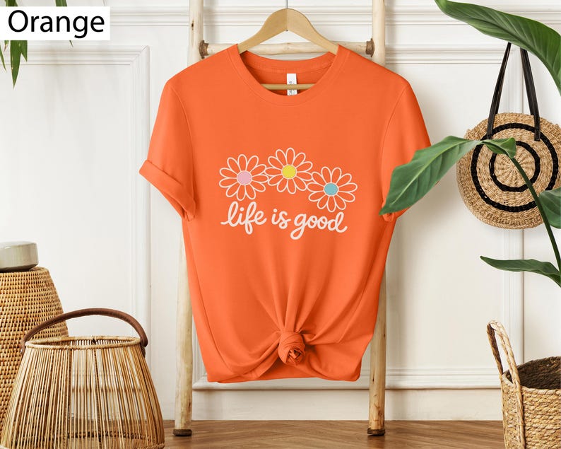 May include: Bright orange t-shirt featuring a floral design and the words "life is good" in white script. The t-shirt is knotted at the waist and hangs on a wooden hanger.