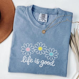 May include: A blue Comfort Colors t-shirt featuring a daisy design and the words "life is good" in white script. A gold necklace with a small pendant is on the shirt. The shirt is folded and has a vintage appearance.