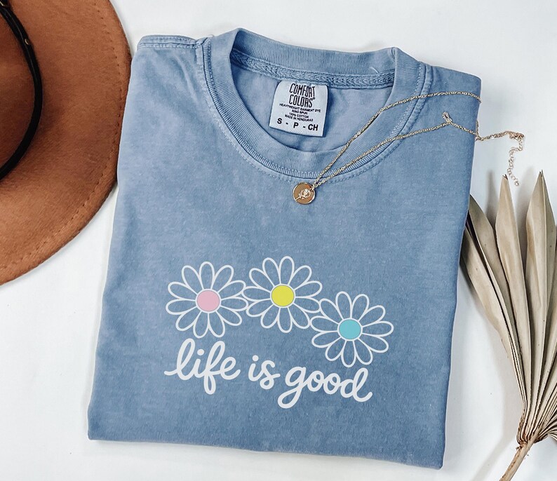 May include: A blue Comfort Colors t-shirt featuring a daisy design and the words "life is good" in white script. A gold necklace with a small pendant is on the shirt. The shirt is folded and has a vintage appearance.