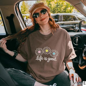 May include: Espresso-coloured t-shirt featuring the phrase "life is good" and three daisy graphics. The shirt has a relaxed fit and is being worn in a car. The shirt is a solid colour with a crew neck.