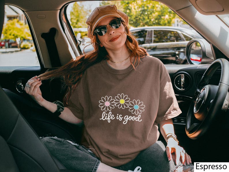 May include: Espresso-coloured t-shirt featuring the phrase "life is good" and three daisy graphics. The shirt has a relaxed fit and is being worn in a car. The shirt is a solid colour with a crew neck.