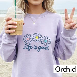 May include: Lilac sweatshirt featuring a daisy design with pink, yellow, and blue centres, and the words "life is good". The sweatshirt has a crew neck and long sleeves. The word "Orchid" is printed at the bottom.