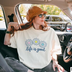 May include: Cream-coloured t-shirt featuring a daisy graphic and the words "life is good" in a script font. The daisies have pink, yellow, and blue centres. The shirt is worn by a person in a car, wearing a brown hat and sunglasses.