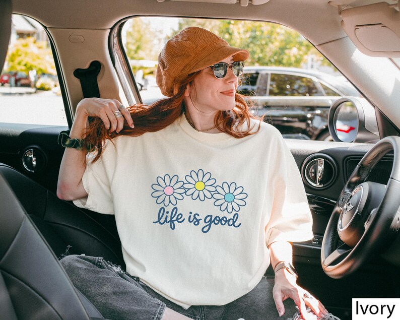 May include: Cream-coloured t-shirt featuring a daisy graphic and the words "life is good" in a script font. The daisies have pink, yellow, and blue centres. The shirt is worn by a person in a car, wearing a brown hat and sunglasses.