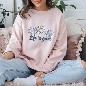 May include: A pale pink sweatshirt featuring a daisy design and the words "life is good". The daisy graphic has pink, yellow, and blue centres. The sweatshirt is a casual, long-sleeved top, suitable for everyday wear.